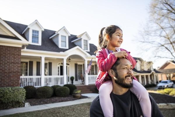 un père avec sa fille sur les épaules devant leur maison