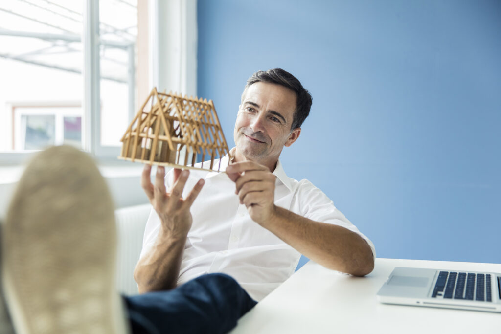 bien choisir sa SCPI : ​​Un homme, les pieds posés sur son bureau. Il tient entre ses mains une maison à l’état de maquette.​