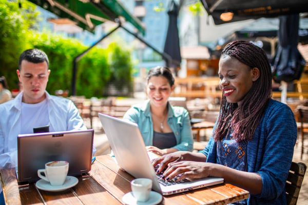 Des amis assis à une table d’une terrasse ensoleillée consultent leurs ordinateurs portables.